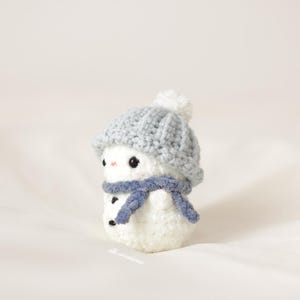 Puede incluir: Un pequeño muñeco de nieve de crochet blanco que lleva un gorro de crochet gris con un pompón blanco y una bufanda azul.