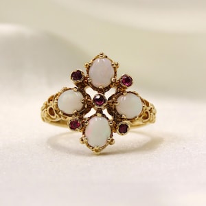 Könnte beinhalten: Ein goldener Ring mit einem kunstvollen Design, besetzt mit fünf ovalen, weißen Opal-Edelsteinen und mehreren kleinen, runden, roten Rubin-Edelsteinen. Der Ring hat ein detailliertes Band im Filigranstil.