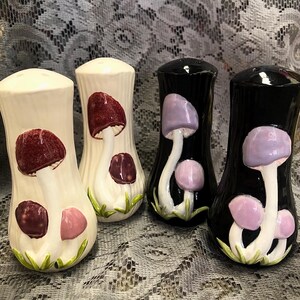 May include: Four ceramic salt and pepper shakers with mushroom designs. Two shakers are white with burgundy and pink mushrooms, and two are black with purple and pink mushrooms. The shakers have a glossy finish and a whimsical aesthetic.