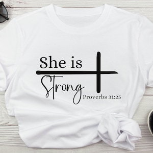 Może przedstawiać: Biała koszulka z czarnym napisem "She is Strong" z krzyżem i tekstem "Proverbs 31:25" poniżej.