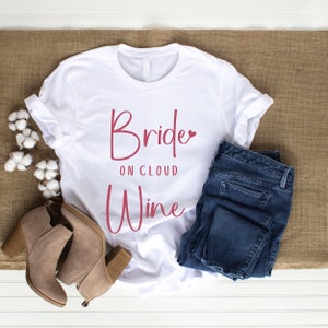 Op de afbeelding: Een wit t-shirt met de tekst "Bride on Cloud Wine" in een bordeauxrode scriptletter. Het shirt wordt gecombineerd met een blauwe jeans en bruin suède enkellaarsjes. Katoentakken zijn aan de zijkant geplaatst, wat een casual bruidsthema creëert.