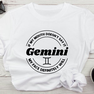 May include: White t-shirt with a black graphic design featuring the word "Gemini" in a bold font, encircled by the text "If my mouth doesn't say it, my face definitely will." The Gemini zodiac symbol is below the word.