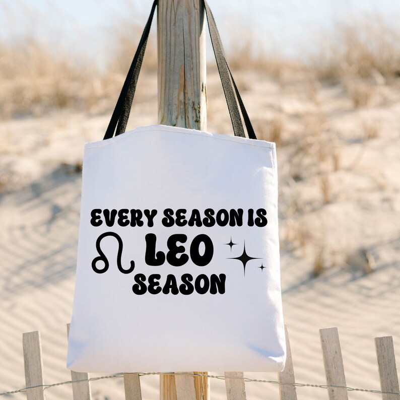 May include: White tote bag with black text that reads "Every season is ♌ Leo season". The bag is hanging from a wooden post against a sandy background.