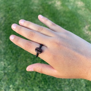 May include: A black ring with a coffee bean design. The ring features a dark brown coffee bean at the center, set on a black band. The ring is shown on a hand against a green background.