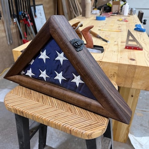 Puede incluir: Una caja triangular de madera con un frente de vidrio, que muestra una bandera estadounidense doblada. La caja tiene un tinte marrón oscuro y un cierre de metal.