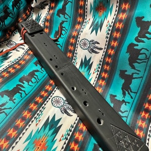 May include: A black, rectangular flute with a decorative geometric design on one end. The flute has several holes and is connected with a leather strap. The flute rests on a turquoise and white patterned fabric with horses and dream catchers.