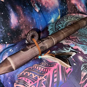 Puede incluir: Una flauta de estilo nativo americano marrón y gris con una correa de cuero y detalles de plumas. La flauta descansa sobre una tela estampada colorida con un diseño de galaxia y plumas. Un artículo decorativo.