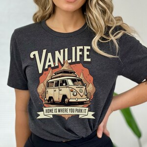 May include: A dark gray t-shirt with a vintage-style illustration of a white van with a roof rack, driving through a mountain landscape. The text "VANLIFE" is in a large, stylized font above the van, and the text "HOME IS WHERE YOU PARK IT" is below the van.