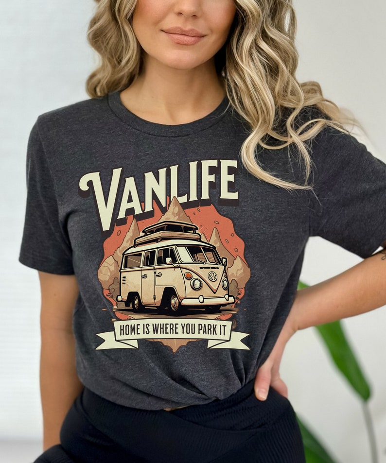 May include: A dark gray t-shirt with a vintage-style illustration of a white van with a roof rack, driving through a mountain landscape. The text "VANLIFE" is in a large, stylized font above the van, and the text "HOME IS WHERE YOU PARK IT" is below the van.
