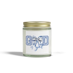 May include: A glass jar candle with a gold lid. The label features the words "GOD is good" in blue floral print. The candle is a decorative item.