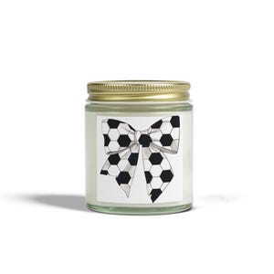 May include: A glass jar candle with a gold lid. The label features a black and white soccer ball bow design. The candle is a decorative item, perfect for home decor or as a gift. The jar is filled with a white wax.