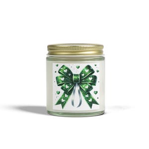 May include: A candle in a glass jar with a gold lid. The label features a green bow with white heart accents and silver ribbons. Small green hearts are scattered around the bow design. The candle is likely scented.