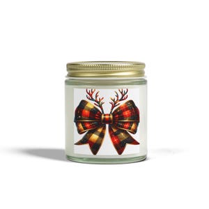 May include: A glass jar candle with a gold lid. The label features a plaid bow with antlers in red, black, and gold. The candle is a festive item, suitable for holiday decor or gifting.