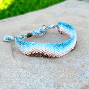 May include: A handmade bracelet with a woven design. The bracelet features a gradient of colors, including shades of blue, white, tan, and brown. The bracelet is resting on a stone surface.