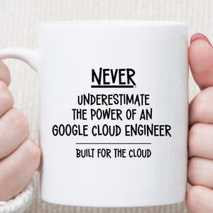 May include: A white ceramic mug featuring black printed text that reads: NEVER UNDERESTIMATE THE POWER OF AN GOOGLE CLOUD ENGINEER, with the phrase BUILT FOR THE CLOUD printed below. The mug has a smooth finish and a standard handle on the left side.