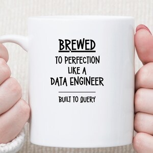 May include: A white ceramic mug featuring black text that reads BREWED TO PERFECTION LIKE A DATA ENGINEER and BUILT TO QUERY. The mug has a smooth finish and a curved handle on the left side, presented against a neutral, textured background.