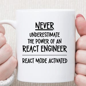 May include: White ceramic mug with the text "NEVER UNDERESTIMATE THE POWER OF AN REACT ENGINEER" in black. Below the text is "REACT MODE ACTIVATED". The mug is held by a person wearing a white sweater.