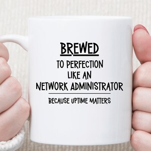 May include: A white ceramic mug featuring black text that reads: BREWED TO PERFECTION LIKE AN NETWORK ADMINISTRATOR BECAUSE UPTIME MATTERS. The mug has a smooth finish and a curved handle on the left side, designed for holding hot beverages.