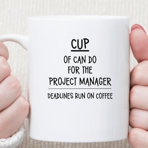 May include: A white ceramic mug featuring black text that reads CUP OF CAN DO FOR THE PROJECT MANAGER and DEADLINES RUN ON COFFEE. The mug has a curved handle on the left side and a smooth, cylindrical body with a clean, minimalist design.