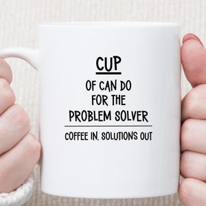 May include: A white ceramic mug featuring black text that reads CUP OF CAN DO FOR THE PROBLEM SOLVER with the additional line COFFEE IN, SOLUTIONS OUT printed below. The mug has a smooth finish and a curved handle on the left side for easy gripping.