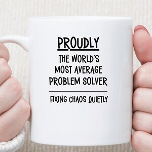 May include: A white ceramic mug featuring black text that reads: PROUDLY THE WORLD'S MOST AVERAGE PROBLEM SOLVER FIXING CHAOS QUIETLY. The mug has a smooth finish and a curved handle on the left side, presented against a neutral, soft-textured background.