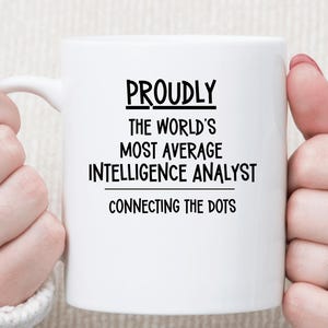 May include: A white ceramic mug featuring black printed text that reads PROUDLY THE WORLD'S MOST AVERAGE INTELLIGENCE ANALYST CONNECTING THE DOTS. The mug has a smooth finish and a curved handle, presented in a clear, centered view for easy reading.