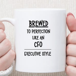 May include: White ceramic mug with the text "BREWED TO PERFECTION LIKE AN CFO EXECUTIVE STYLE" in black. The mug is held by two hands with rings on the fingers.