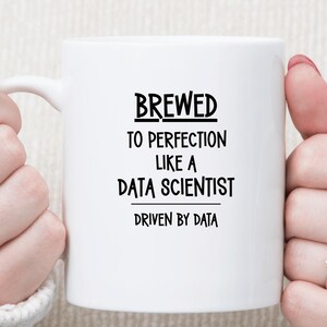 May include: A white ceramic mug featuring black text that reads BREWED TO PERFECTION LIKE A DATA SCIENTIST and DRIVEN BY DATA. The mug has a smooth finish and a curved handle on the left side, presented against a neutral, textured background.