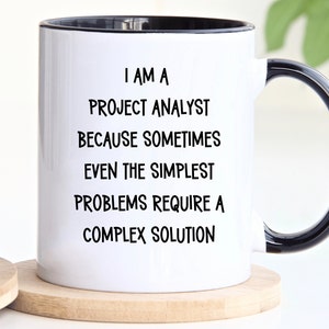 May include: White ceramic mug with a black rim and handle. The mug has black text that reads "I am a Project Analyst because sometimes even the simplest problems require a complex solution."