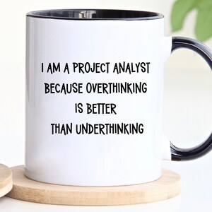 May include: White ceramic mug with a black rim and handle. The mug has black text that reads "I am a project analyst because overthinking is better than underthinking."