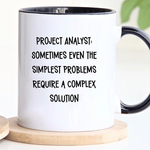 May include: White ceramic mug with a black rim and handle. The mug has black text that reads "Project Analyst: Sometimes even the simplest problems require a complex solution."