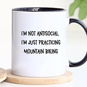 May include: White ceramic mug with a black rim and handle. The mug has black text that reads "I'm not antisocial, I'm just practicing mountain biking."