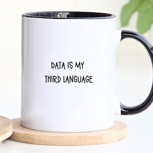 May include: White ceramic mug with a black rim and handle. The mug has the text "DATA IS MY THIRD LANGUAGE" printed on it.