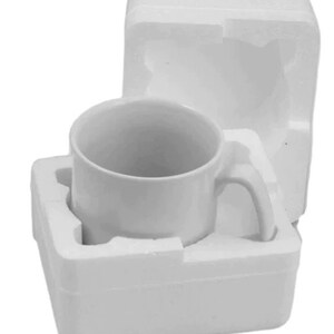 K&ouml;nnte beinhalten: Eine wei&szlig;e Keramiktasse mit gebogenem Henkel, verpackt in wei&szlig;em Schaumstoff. Die Tasse ist zylindrisch und leer. Die Verpackung dient zum Schutz der Tasse w&auml;hrend des Transports und der Lagerung.