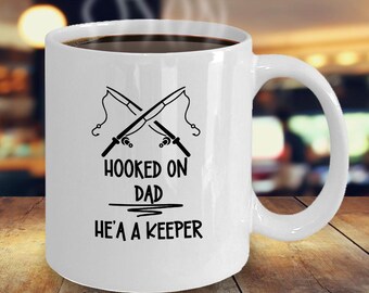 May include: White ceramic coffee mug with a black design of two crossed fishing rods and the text "Hooked on Dad He's a Keeper".