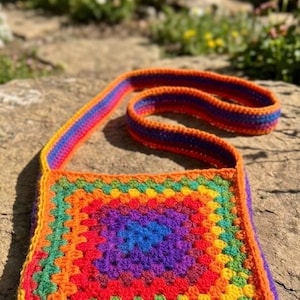 May include: A handmade, colorful crochet bag with a rainbow pattern. The square bag features a vibrant spectrum of colors, including red, orange, yellow, green, blue, and purple. The strap is also rainbow-colored, creating a cohesive and eye-catching design.