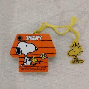 May include: A yellow and orange plastic keychain featuring Snoopy, the beagle from the Peanuts comic strip, sitting in his doghouse. The keychain has a yellow string attached to it and a small plastic figure of Woodstock, Snoopy's best friend, hanging from the string. The keychain has the text "Snoopy" printed on it and the copyright information "PEANUTS Characters. Copr. © 1958. 1965 United Feature Syndicate, Inc."