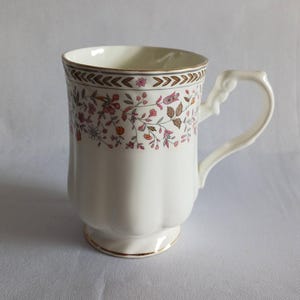 May include: White ceramic mug with a floral design in pink, brown, and gold. The mug has a decorative handle and a gold rim. The floral pattern wraps around the top of the mug.