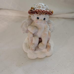 May include: A small, off-white angel figurine with a halo of dried flowers and a cloud base. The angel has small wings and is seated, holding a staff. The figurine has a slightly aged appearance.