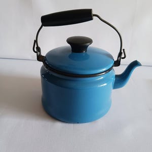 May include: A small, blue enamel teapot with a black handle and lid. The teapot has a curved spout and a wire handle that arches over the top. The lid is also black, and the body of the teapot is a solid, light blue color.