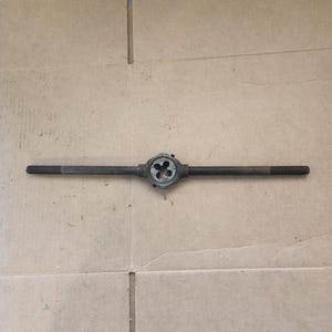 May include: A metal pipe threader tool with a black finish. The tool has a handle on each end and a die in the center.