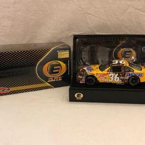 Op de afbeelding: Een geel en bruin model van een raceauto met nummer 36 en Snickers-branding, gepresenteerd in een zwarte doos. Daarnaast een zwart en goudkleurig patroon doos met het Elite-logo.