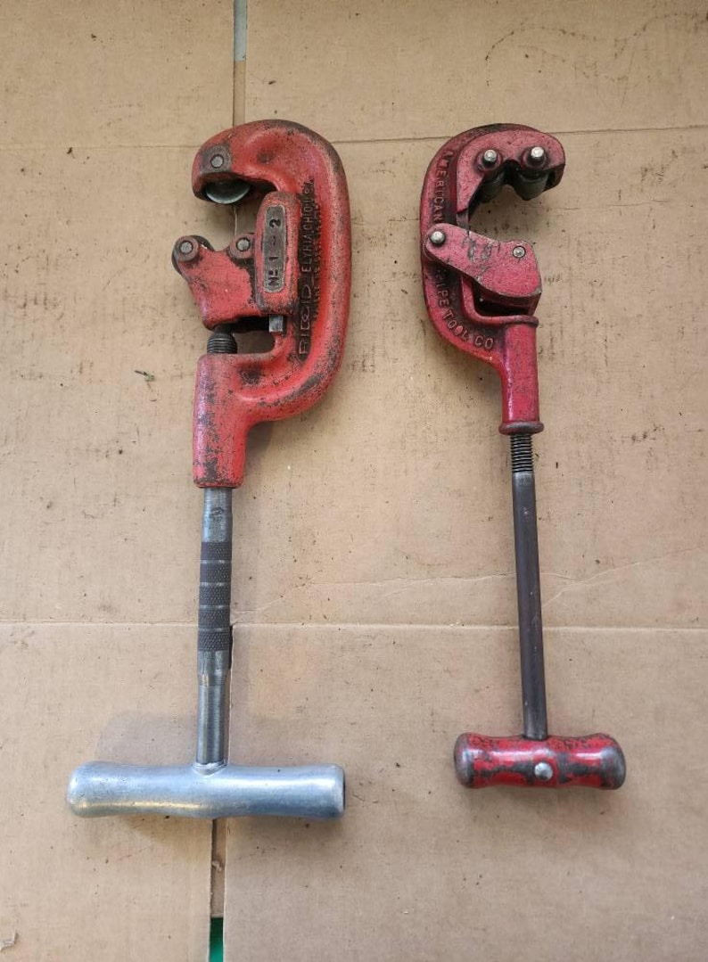 K&ouml;nnte beinhalten: Zwei rote Rohrschneider, einer mit einem silbernen Griff und der andere mit einem roten Griff. Der Rohrschneider mit dem silbernen Griff hat den Text "RIDGID  ELECTRIC  MFG. CO.  NO. 1-2" an der Seite. Der Rohrschneider mit dem roten Griff hat den Text "AMERICAN PIPE TOOL CO." an der Seite.