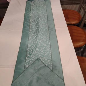May include: A rectangular, teal-colored table runner with a floral pattern in the center. The runner has a teal border and is displayed on a white tablecloth. The runner is approximately 60 inches long.