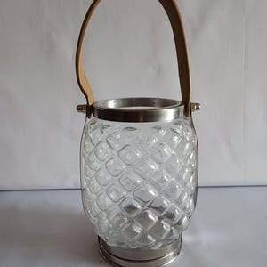 May include: Clear glass lantern with a textured, diamond-like pattern. It has a silver-colored metal rim and base, and a tan leather handle. The lantern is designed to hold a candle or other light source.