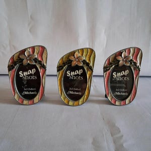 May include: Three decorative photo frames with a floral design. Each frame has a curved top with a flower accent and the text "Snap shots". The frames are 2x3 inches (5x8cm) and have red, yellow, and cream stripes.