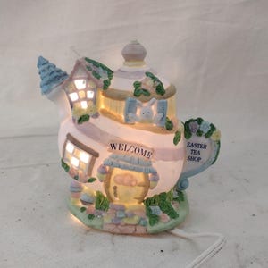 May include: A ceramic Easter tea shop shaped like a teapot, with a light inside. The teapot is painted in pastel colors and features a small house, a bunny, and the words "WELCOME" and "EASTER TEA SHOP".