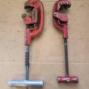 K&ouml;nnte beinhalten: Zwei rote Rohrschneider, einer mit einem silbernen Griff und der andere mit einem roten Griff. Der Rohrschneider mit dem silbernen Griff hat den Text "RIDGID  ELECTRIC  MFG. CO.  NO. 1-2" an der Seite. Der Rohrschneider mit dem roten Griff hat den Text "AMERICAN PIPE TOOL CO." an der Seite.