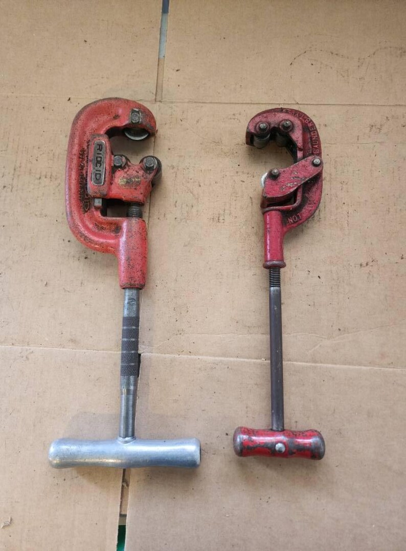 K&ouml;nnte beinhalten: Zwei rote Rohrschneider mit silbernen Griffen. Der Schneider links hat den Schriftzug "RIDGID" aufgedruckt. Der Schneider rechts hat den Schriftzug "BAUMDER" aufgedruckt.