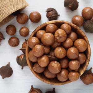 以下が含まれることがあります： マカダミアナッツがいっぱい入った木製のボウル。周りにはナッツが散らばっています。ナッツは薄茶色で、滑らかな質感です。ジュートの袋が左上に一部見えています。